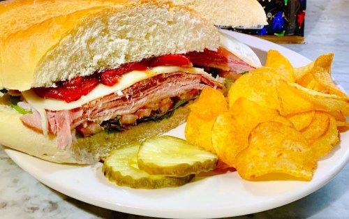 A hearty sandwich with layers of meat, cheese, and toppings, served with potato chips and pickles on a white plate.