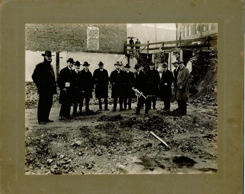 A group of men in early 20th-century attire stands on a construction site, examining the ground.