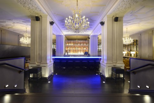 Elegant bar interior with a blue-lit counter, chandeliers, and decorative columns, creating a sophisticated ambiance.