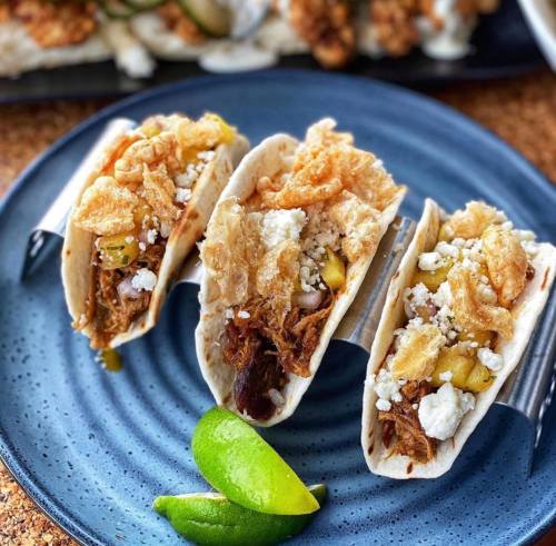Three tacos filled with meat, topped with crispy bits and cheese, served on a blue plate with lime wedges.