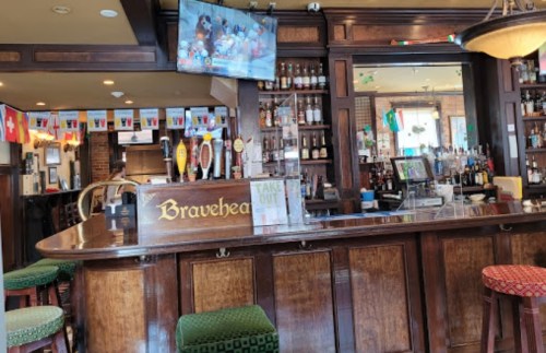 A cozy bar with wooden decor, a TV showing sports, and flags hanging from the ceiling. Bar stools are visible.