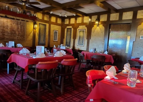 Cozy restaurant interior with red tablecloths, wooden chairs, and warm lighting, featuring neatly set tables.