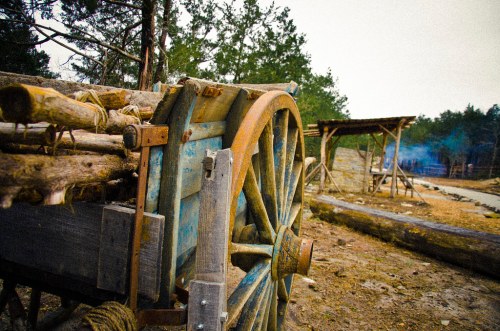A close-up of a weathered wooden cart wheel, with a rustic landscape and structures in the background.