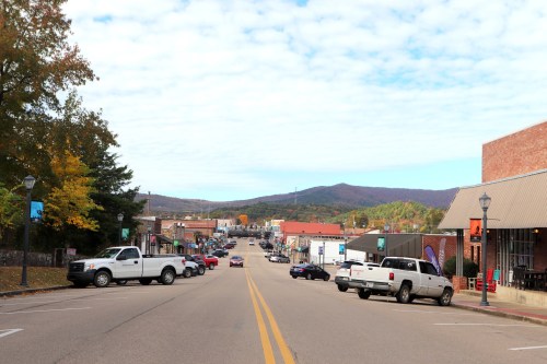 A small town street lined with shops and parked cars, with mountains in the background under a cloudy sky.