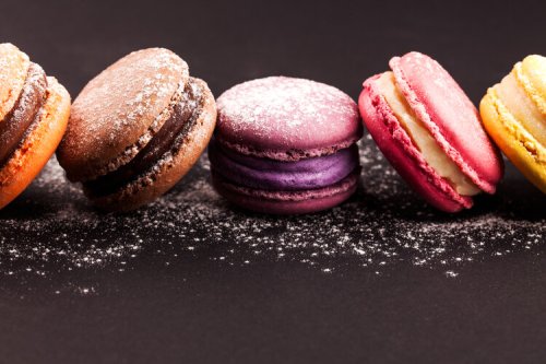 A colorful assortment of macarons in various flavors, dusted with powdered sugar, arranged on a dark background.