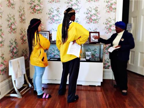 Two visitors in yellow jackets listen to a guide in a blue hat, surrounded by floral wallpaper and historical displays.