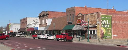 A row of historic brick buildings along a street, featuring shops and parked cars under a clear blue sky.