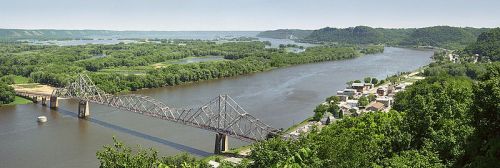 A panoramic view of a river with a bridge, lush greenery, and a small town along the riverbank under a clear sky.