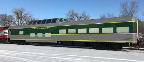 A vintage train car with a green and gray exterior, labeled "Adirondack," parked on a snowy landscape.