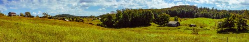A panoramic view of rolling green hills, scattered trees, and rustic buildings under a bright blue sky with fluffy clouds.