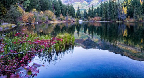 A serene lake surrounded by colorful autumn foliage and evergreen trees, reflecting the mountains in the background.