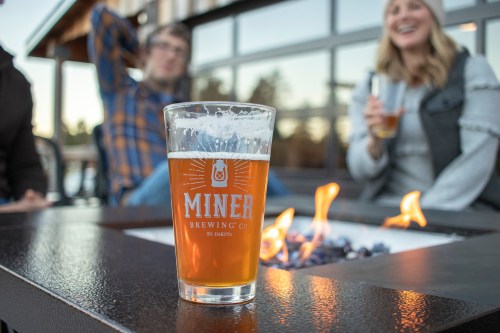 A glass of beer sits on a table with a fire pit, while people enjoy drinks and conversation in the background.