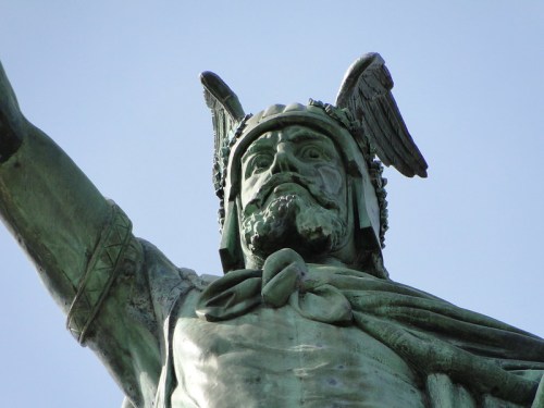 A close-up of a bronze statue of a warrior with a winged helmet, raising one arm against a clear blue sky.