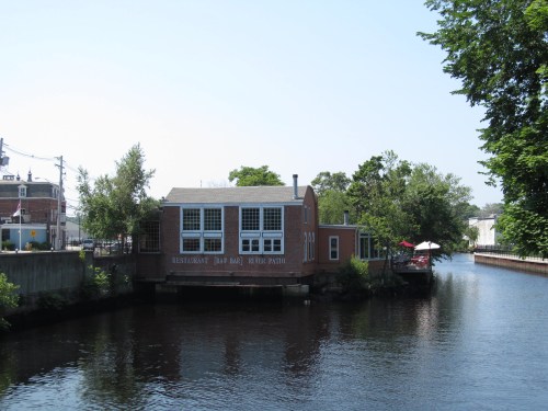 A riverside restaurant with a patio, surrounded by trees and water, on a clear sunny day.