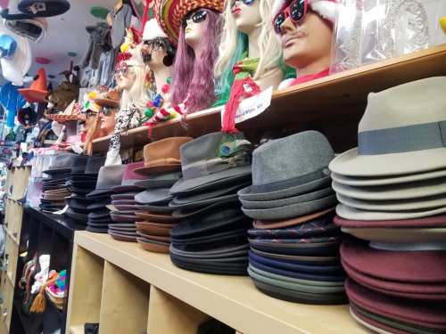 A display of various hats stacked on a shelf, with colorful wigs and accessories in the background.