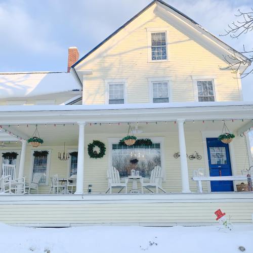 A charming house with a porch, rocking chairs, and holiday decorations, surrounded by fresh snow under a clear sky.