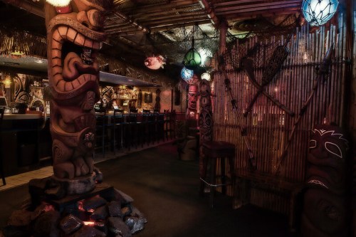 A dimly lit tiki bar featuring carved wooden totems, bamboo decor, and colorful hanging lights.