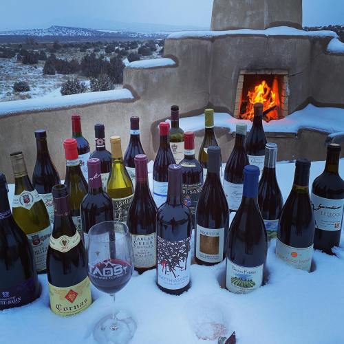 A snowy outdoor setting with a variety of wine bottles arranged in front of a warm fireplace.