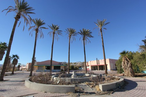 Abandoned plaza with palm trees and a dry fountain, surrounded by derelict buildings under a clear blue sky.