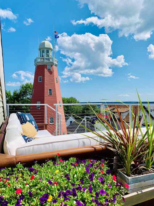 A cozy outdoor seating area with colorful flowers, overlooking a red lighthouse and a bright blue sky with clouds.