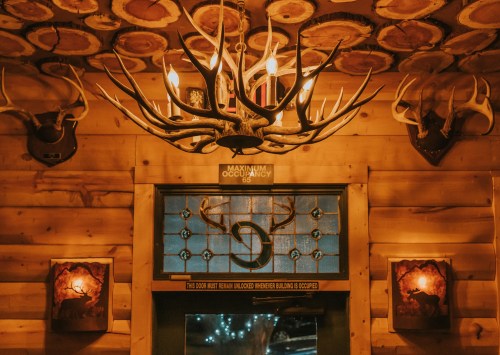 Cozy cabin interior with wooden walls, antler decor, a chandelier, and a stained glass window.