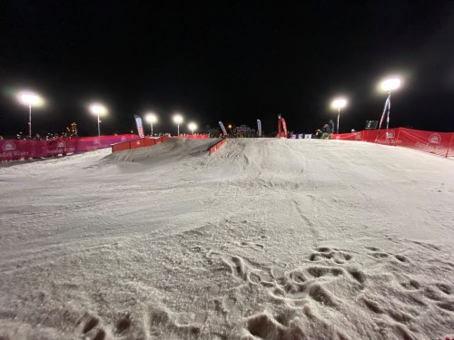 A snowy slope at night, lined with red barriers and illuminated by bright lights, with spectators in the background.
