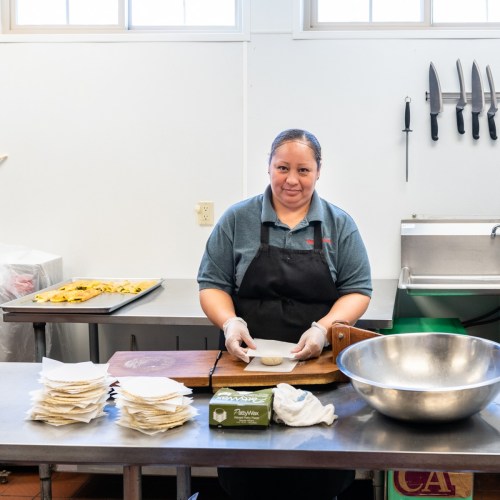 A woman in an apron prepares dough at a kitchen counter, with utensils and food items in the background.