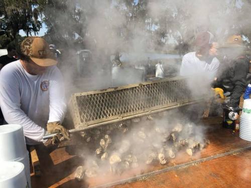 Two people steaming oysters outdoors, surrounded by smoke and a rustic setting, with trees in the background.