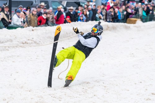 A skier in bright yellow pants falls while being pulled on snow, with a crowd watching in the background.