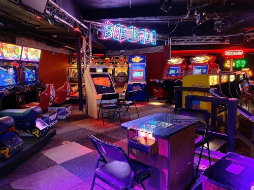 A vibrant arcade featuring various games, including a neon "Skeeball" sign, with empty chairs and colorful machines.