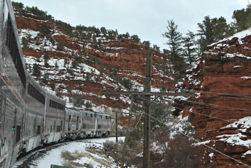 A train curves through a snowy landscape with red rock formations and pine trees in the background.