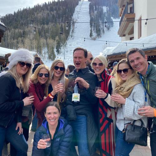 A group of friends poses together outdoors, smiling and holding drinks, with a snowy mountain in the background.