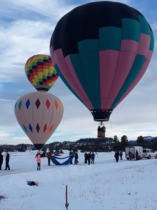 Three colorful hot air balloons are being prepared for flight in a snowy landscape, with people assisting nearby.