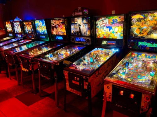 A row of colorful pinball machines illuminated in a dimly lit arcade.