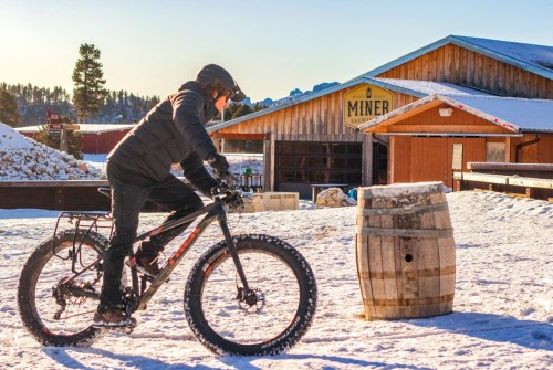 A person rides a fat bike on snow near a wooden building and a barrel, with trees in the background.