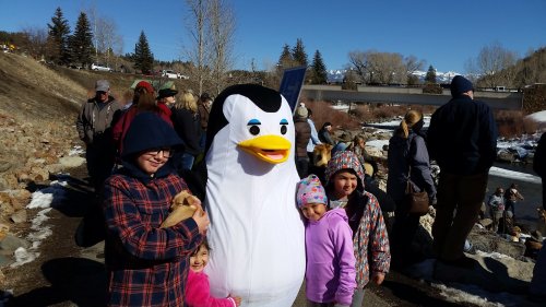A group of children poses with a large penguin mascot at a snowy outdoor event, surrounded by a crowd and trees.