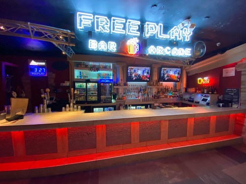 A vibrant bar arcade with neon signs, a well-stocked bar, and multiple screens displaying games.
