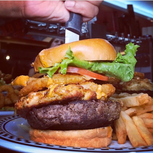 A towering burger with a beef patty, fried chicken, cheese, lettuce, and tomato, served with fries on the side.