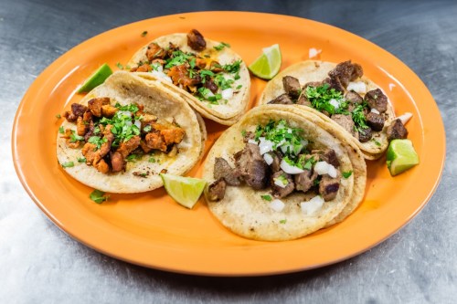 A vibrant orange plate with three tacos filled with different meats, garnished with cilantro and lime wedges.