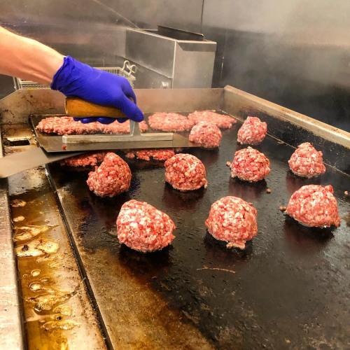 A hand in a blue glove flips raw hamburger patties on a sizzling griddle in a kitchen. Steam rises around the patties.