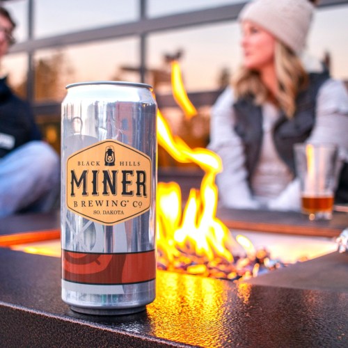 A can of Miner Brewing Co. beer sits near a fire pit, with two people enjoying drinks in the background.
