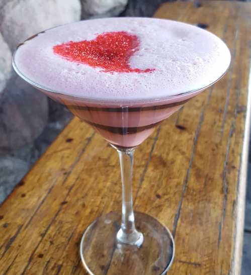 A pink cocktail in a martini glass, topped with a heart-shaped sprinkle of red sugar, on a wooden table.