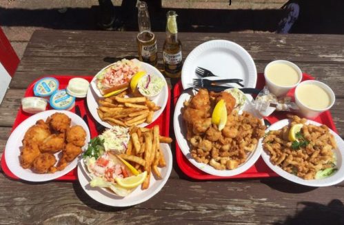 Two trays of seafood dishes with fried fish, calamari, fries, coleslaw, and drinks on a wooden table.