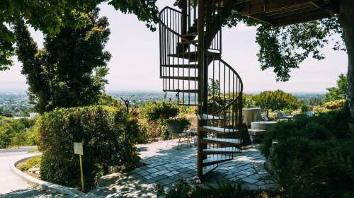 A spiral staircase leads up from a landscaped garden, offering a view of the city below and surrounding greenery.