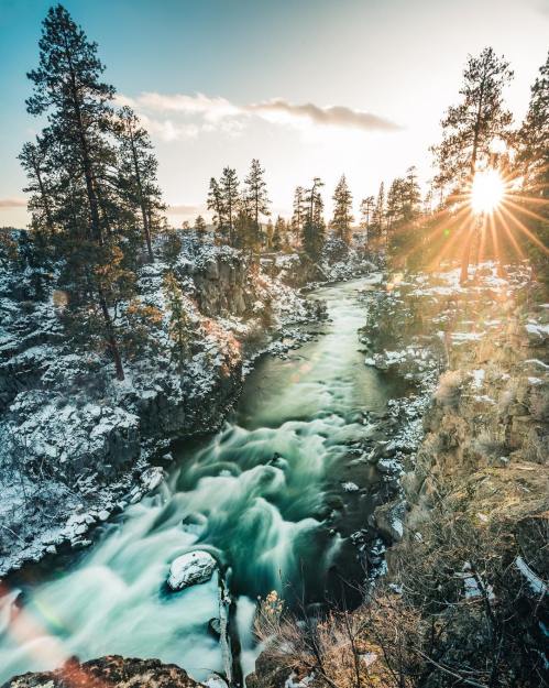 A serene river flows through a snowy landscape, framed by trees and illuminated by a bright sunset.