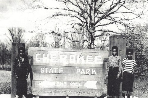 Three children stand beside a sign for Cherokee State Park, with trees in the background and a clear sky.