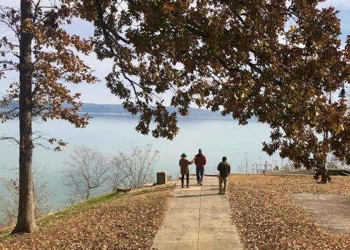 Three people walk along a path by a lake, surrounded by trees with autumn leaves.