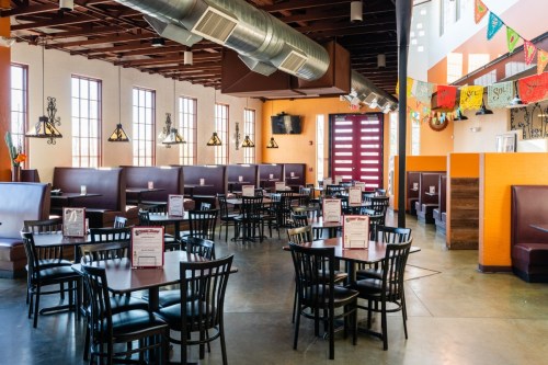 A spacious restaurant interior with wooden tables, black chairs, and colorful decorations hanging from the ceiling.