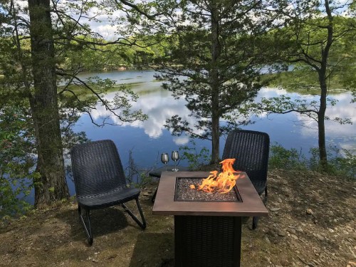 A serene lakeside scene featuring two chairs and a fire pit, surrounded by trees and reflecting water.