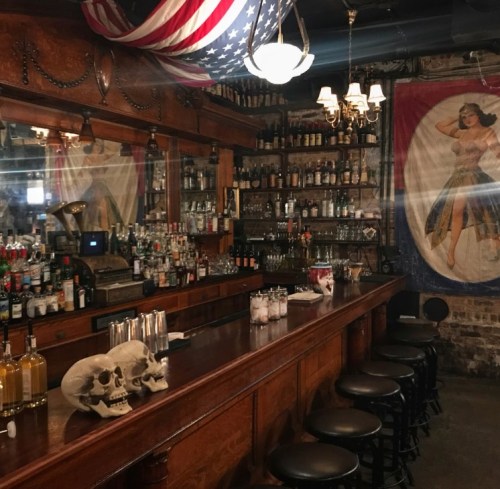 A vintage bar with a wooden counter, shelves of liquor, and a pin-up girl poster, featuring skull decorations.